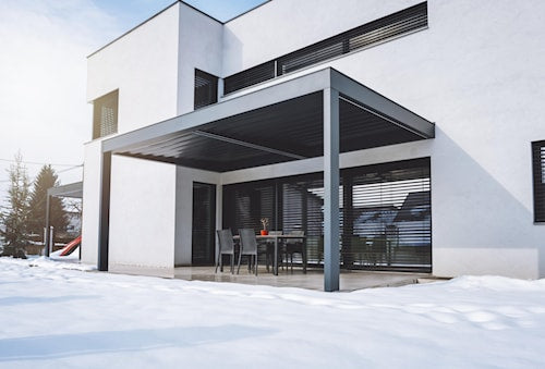 Maison contemporaine blanche avec terrasse couverte en métal gris, table à manger extérieure et sol enneigé reflétant la lumière hivernale.