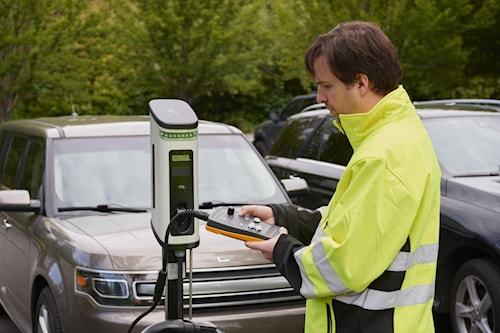 Technicien en veste haute visibilité utilisant un contrôleur portatif pour tester une borne de recharge de véhicule électrique.