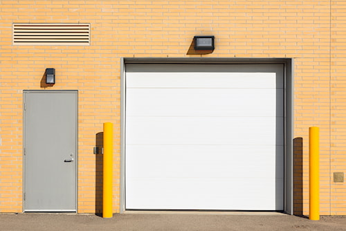 Façade de bâtiment industriel avec porte métallique grise, grande porte de garage blanche, luminaires muraux et bornes de sécurité jaunes