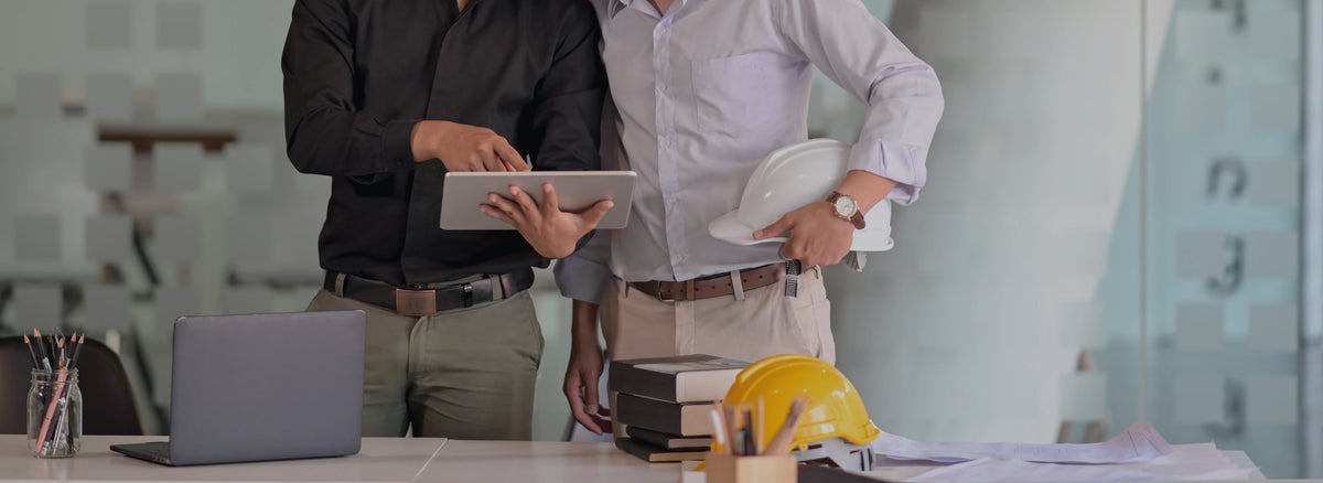 Deux professionnels échangent autour d’une tablette dans un bureau moderne, avec casques de sécurité et plans de chantier sur la table.