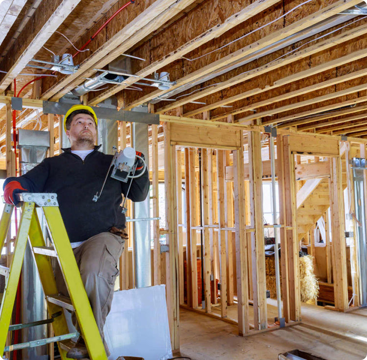 Électricien en casque jaune sur un escabeau dans une maison en construction, tenant des composants électriques, avec conduits apparents au plafond.